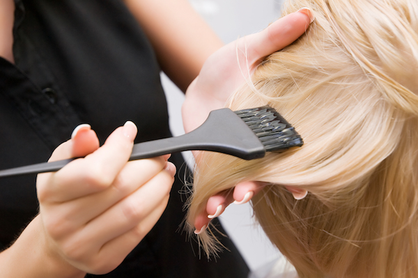 Person trägt mit Pinsel Haarfarbe auf blondes Haar auf und hält dabei eine Strähne mit der Hand fest.
